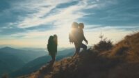 Two hikers in shadow climbing a mountain with the sun behind them.