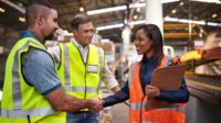 Three workers in safety vests meeting, two shaking hands.