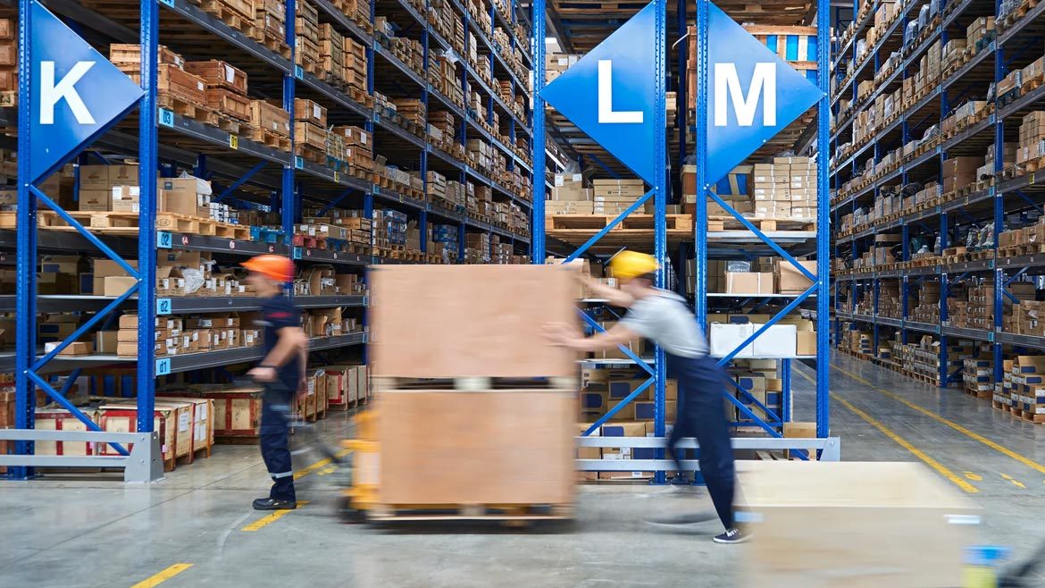 Workers moving stock in a warehouse with lettered blue shelves packed with packages in the background.