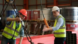 Two construction workers in hard hats and safety vest, using a hoist.