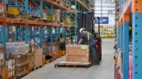 Worker unloading a pallet between large rows of shelves in a supply warehouse.