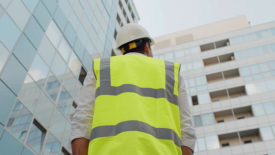 Low angle rear view of engineer with a white safety helmet and safety vest, looking up at modern building outdoors.