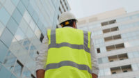 Low angle rear view of engineer with a white safety helmet and safety vest, looking up at modern building outdoors.