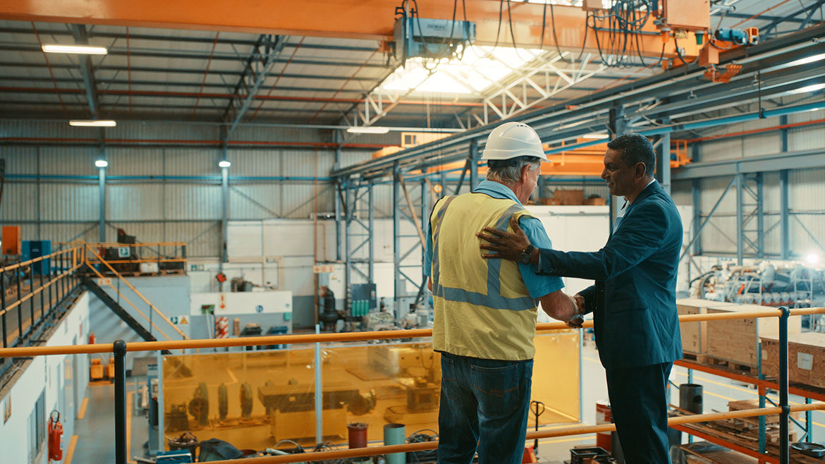 Two men, one in a safety vest and had and another in a business suit, shake hands in agreement while standing on the second level of a shipping warehouse.